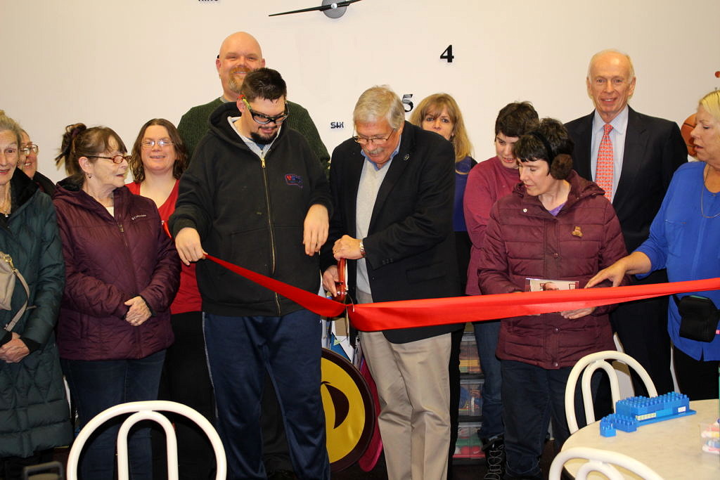 A photo of a large crowd with a man in the middle cutting a red ribbon with oversized scissors