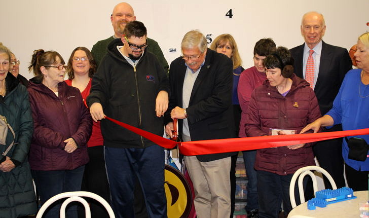A photo of a large crowd with a man in the middle cutting a red ribbon with oversized scissors