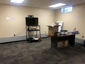 An empty room with bare beige walls, a large TV cart, and a table with paint cans and a large cardboard box on it