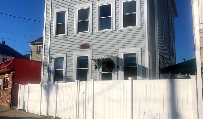 An exterior photo of a gray, three-story residential building behind a white fence