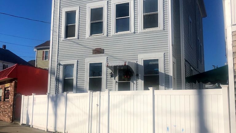 An exterior photo of a gray, three-story residential building behind a white fence