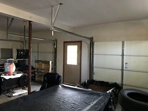 An interior photo of the original garage space with a concrete floor, large garage doors, snow shovels, tires, and other items being stored