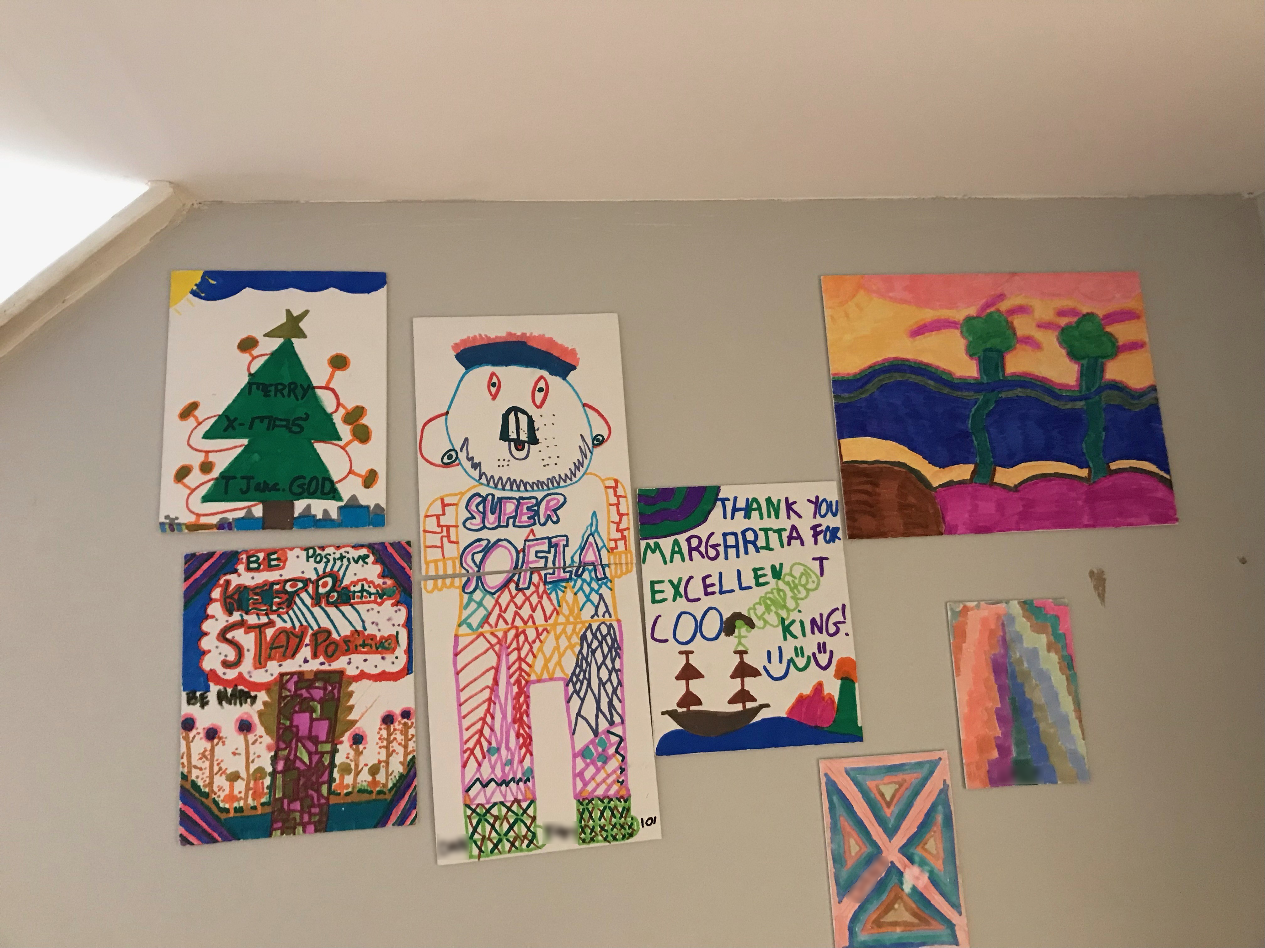 A wall with seven drawings and paintings hanging. The pictures include landscapes, colorful abstract designs, a seascape, a portrait, a Christmas tree, and an image with flowers and the words "Be Positive; Stay Positive"